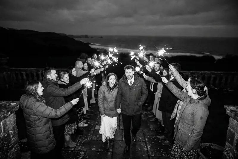 Les mariés traversent de nuit une haie d'invités tenant des cierges magiques sur une terrasse au bord de la mer