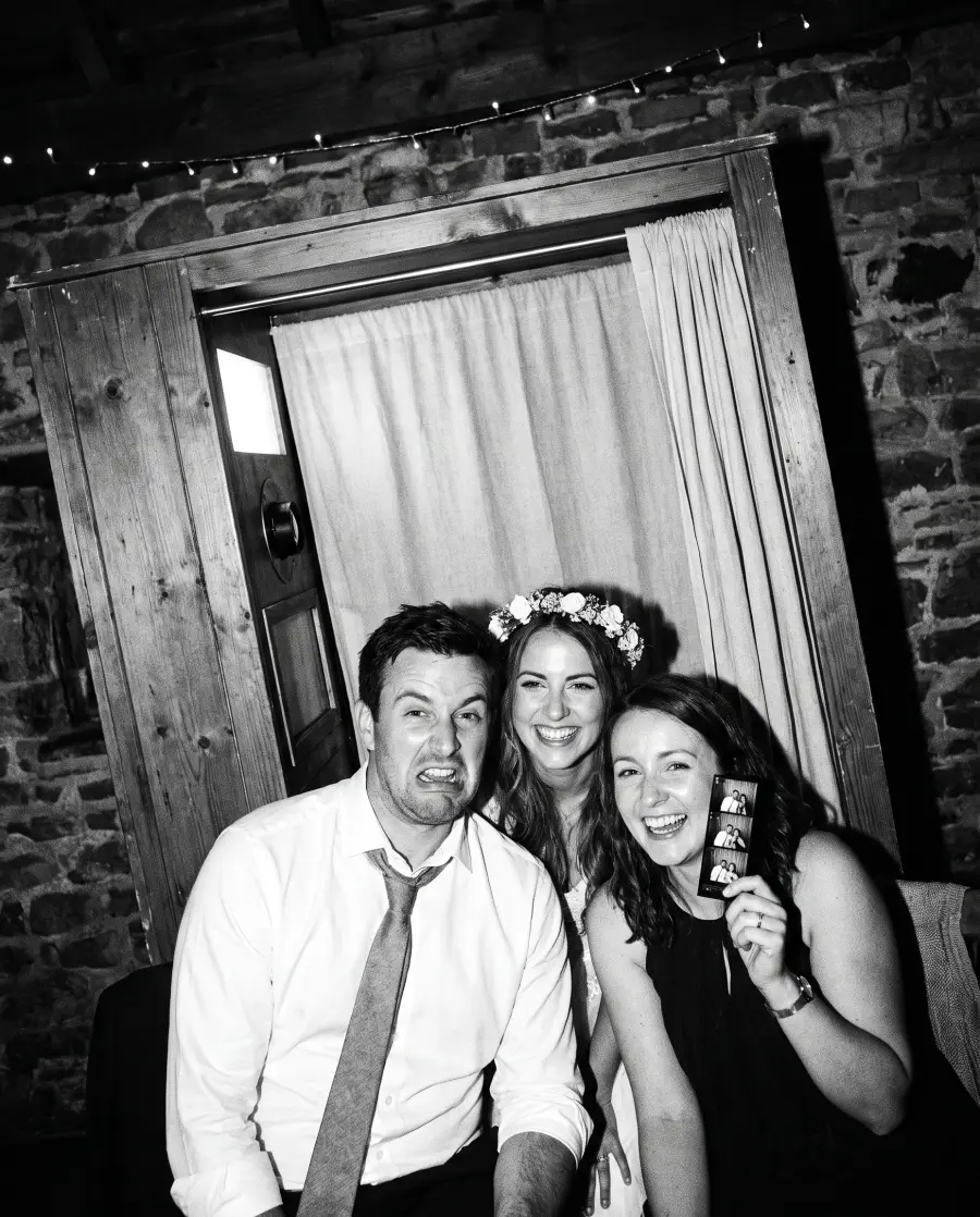 Trois invités de mariage grimacent devant une cabine photo en bois, l'une tient une bande de tirages